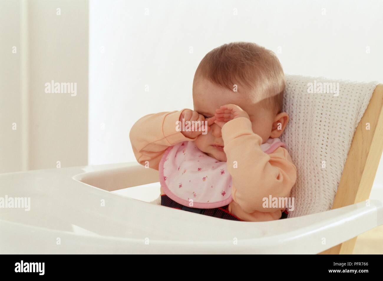 6 Monate alten Baby Mädchen in hoher Stuhl ihre Augen reiben Sitzen Stockfoto
