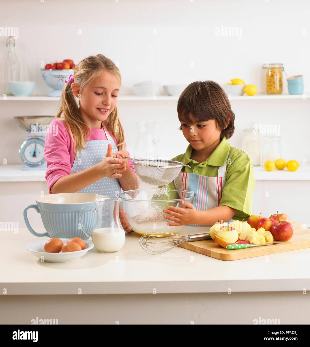 Mehl in Schüssel sieben Mädchen, junge Holding Schüssel, Eier, geschält und in Scheiben geschnittenen Äpfel, Schneebesen, und einen Krug Milch in der Nähe auf der Küchenarbeitsplatte Stockfoto