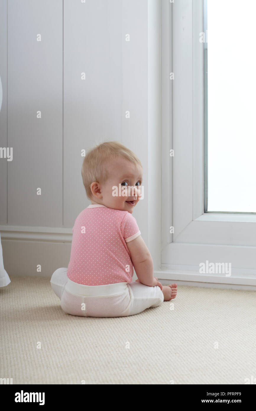 Baby (9 Monate) sitzt auf dem Boden mit dem Rücken zur Kamera gedreht, mit Blick über die Schulter Stockfoto