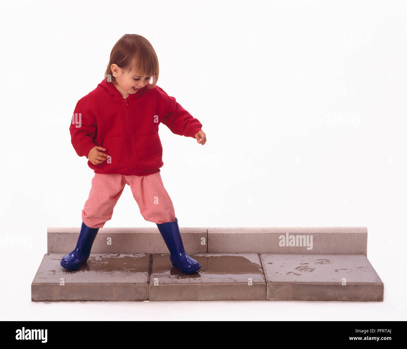 Kleinkind das Tragen der roten Kapuzen Top, pink und blau tousers Gummistiefel Spritzen in Pfütze auf Pflasterstein Stockfoto