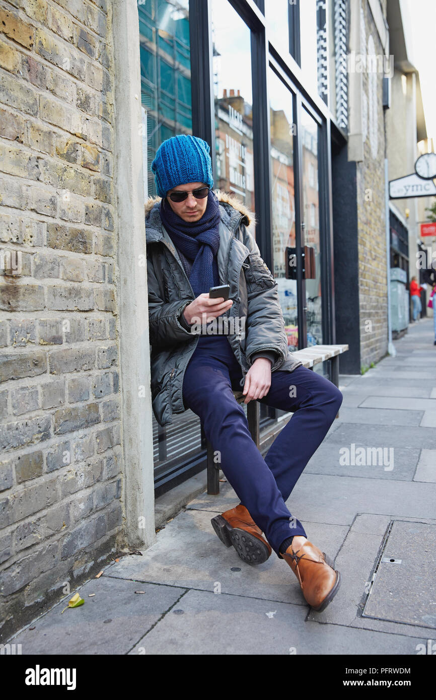 Junger Mann auf einer Bank sitzen außerhalb, auf sein Handy zu schielen. Stockfoto