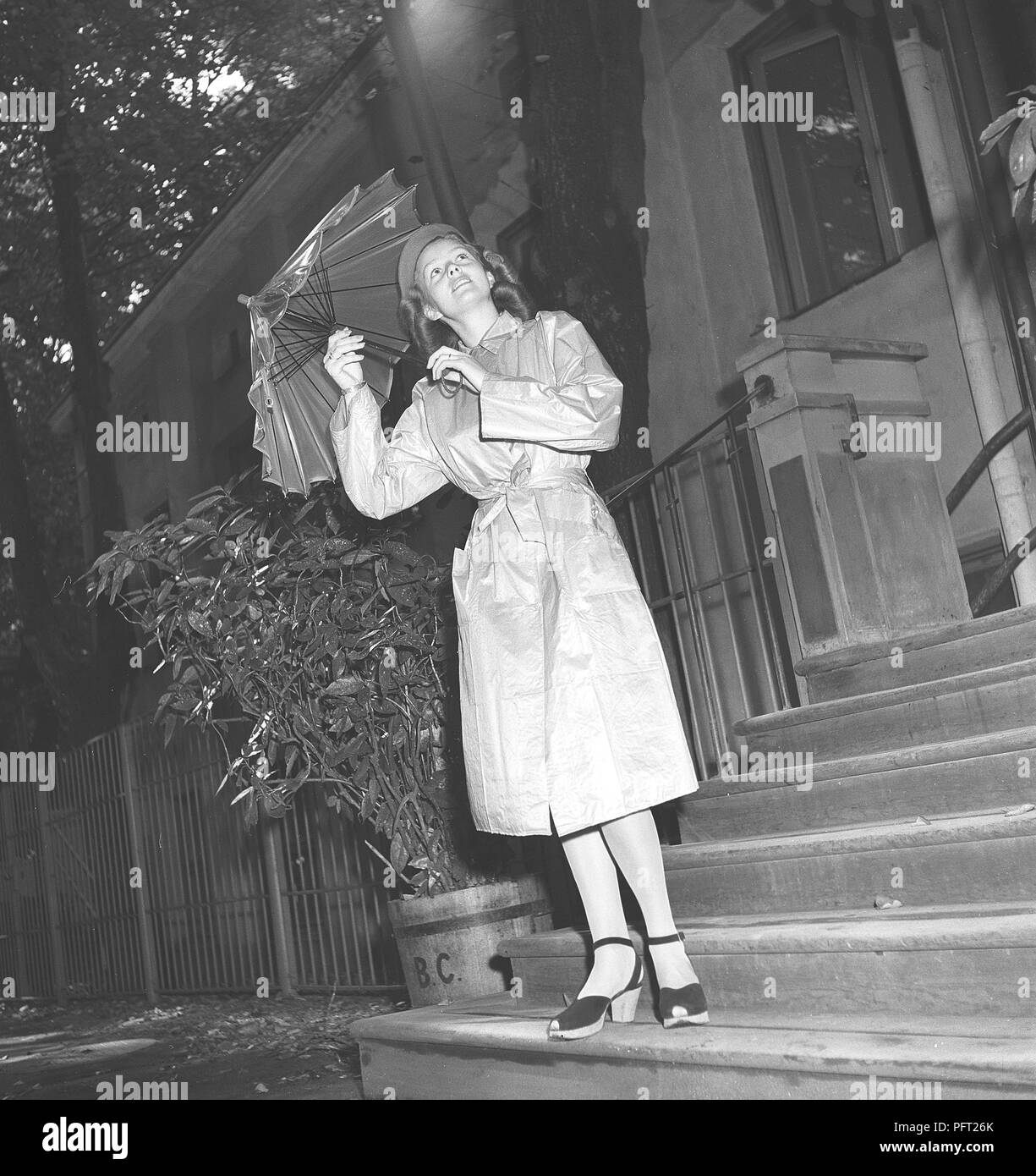 Frau aus den 1940er Jahren im Regenmantel. Eine junge Frau, die einen Regenmantel trägt und einen Schirm hält, schaut in den Himmel, um zu sehen, ob der Regen weiter fließt. Schweden 1946 Ref. V112-5 Stockfoto
