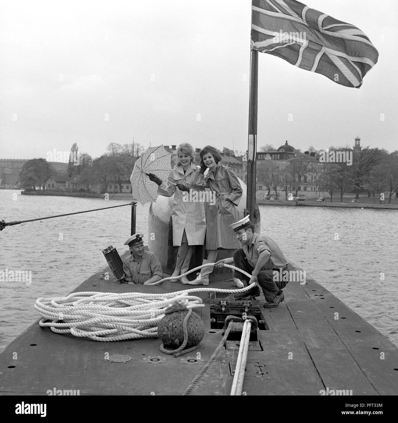 60er Jahre Mode. Zwei fashion Modelle und die Besatzung der Britischen u-boot Alcide. Das U-Boot liegt im Hafen von Stockholm verankert. Die Modelle tragen die herbste Mode Der regenjacken. Schweden Mai 1962 Stockfoto