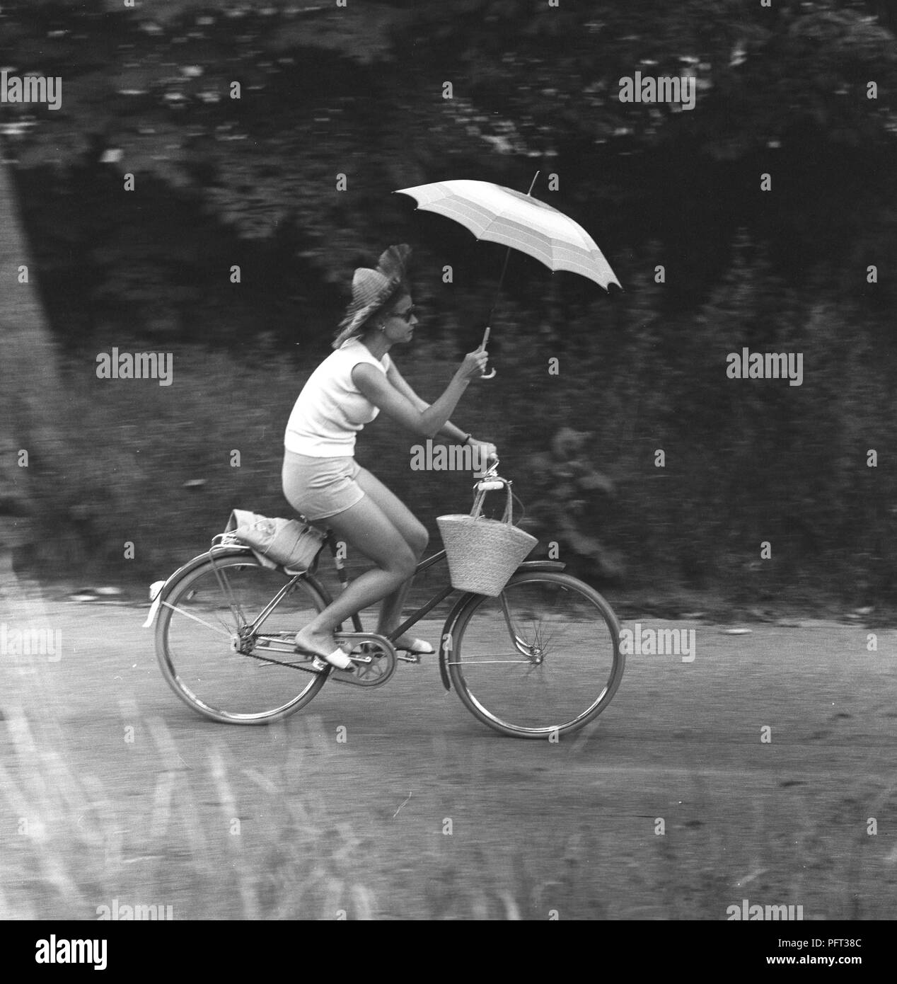 Regen in den 1950er Jahren. Eine junge Frau reitet ein Fahrrad Holding einen Regenschirm zu erhalten zu vermeiden nass vom Regen. Schweden Juli 1959 Stockfoto