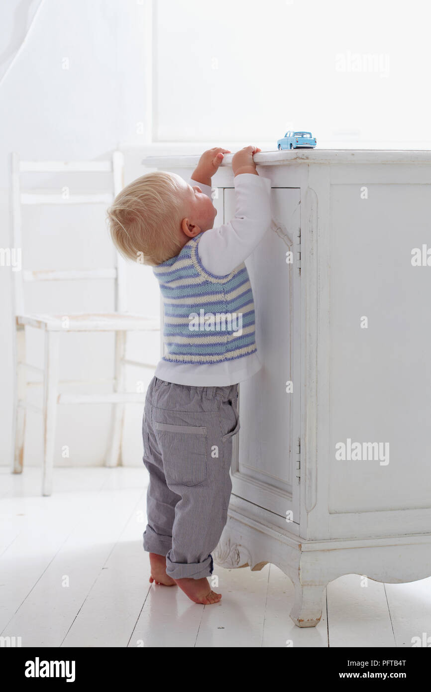 Baby Boy steht auf Zehenspitzen erreichen für blaue Spielzeugauto, 14 Monate Stockfoto
