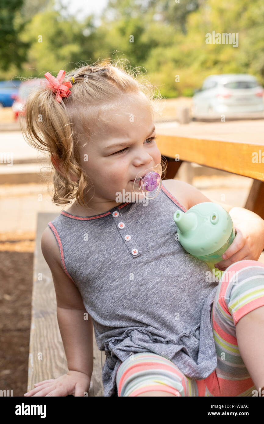 20 Monate altes Mädchen an einem Picknicktisch sitzen, während Ihr sippy cup Holding, suchen sehr in Gedanken verloren Stockfoto