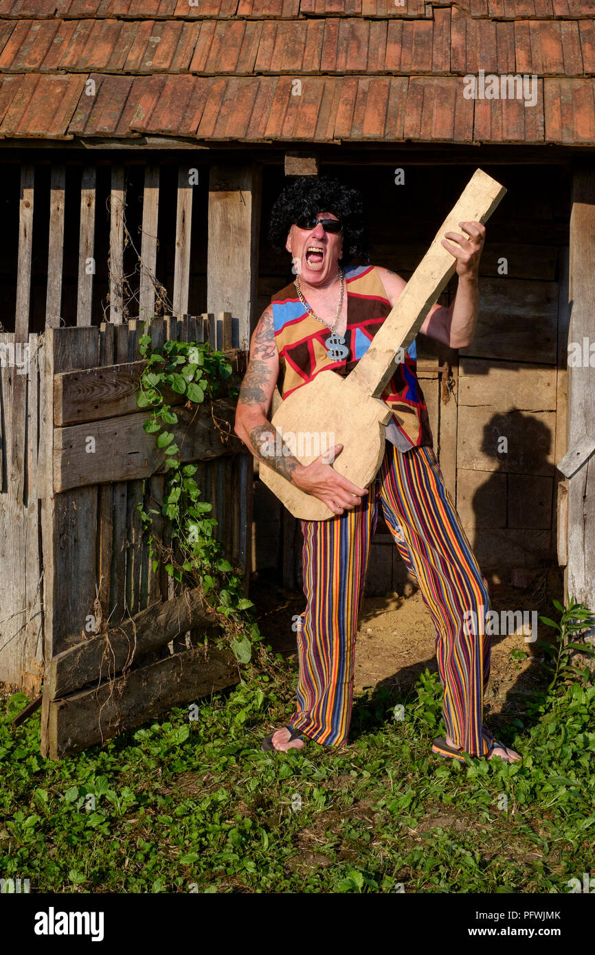 Spoof Portrait von älteren männlichen trägt Perücke Sonnenbrille helle Kleidung und Medaillon während der Wiedergabe eines gefälschten Gitarre Mitte Lebenskrise Konzept Ungarn Stockfoto