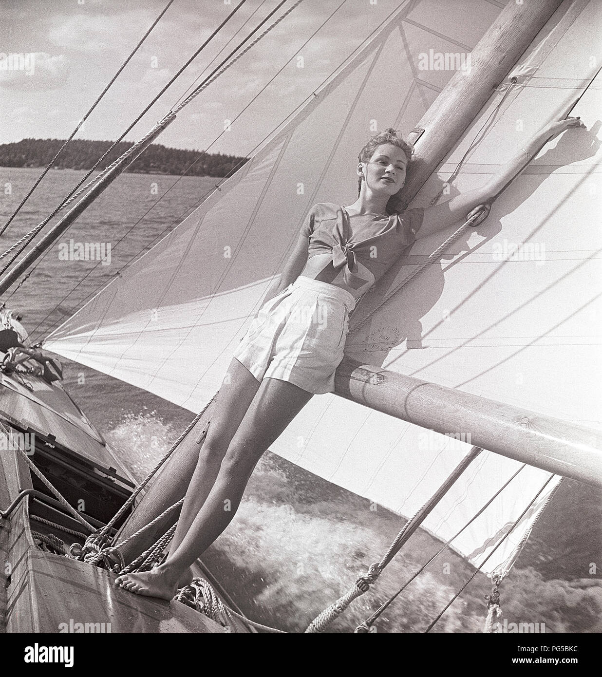 1940 s Segelboot. Eine junge Frau ist an Bord eine modische Segelboot und stehen an Deck und lehnte sich auf den Mast und die Segel, wenn es Kreuzfahrten mit Wind in den Segeln. Sie trägt eine typische 40er Jahre Mode mit einem T-Shirt und Shorts. Schweden 1946 Foto Kristoffersson ref AC 101-2 Stockfoto