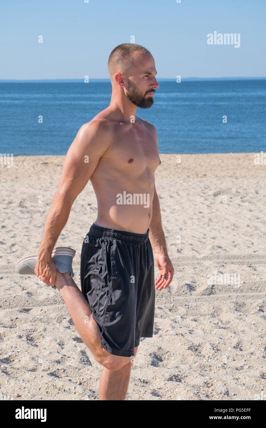 Eine junge attraktive athletischen Mann erstreckt sich Quad beinmuskulatur am Strand Sand Meer Hintergrund nicht tragen Shirt vor der Ausführung der Übung. Stockfoto