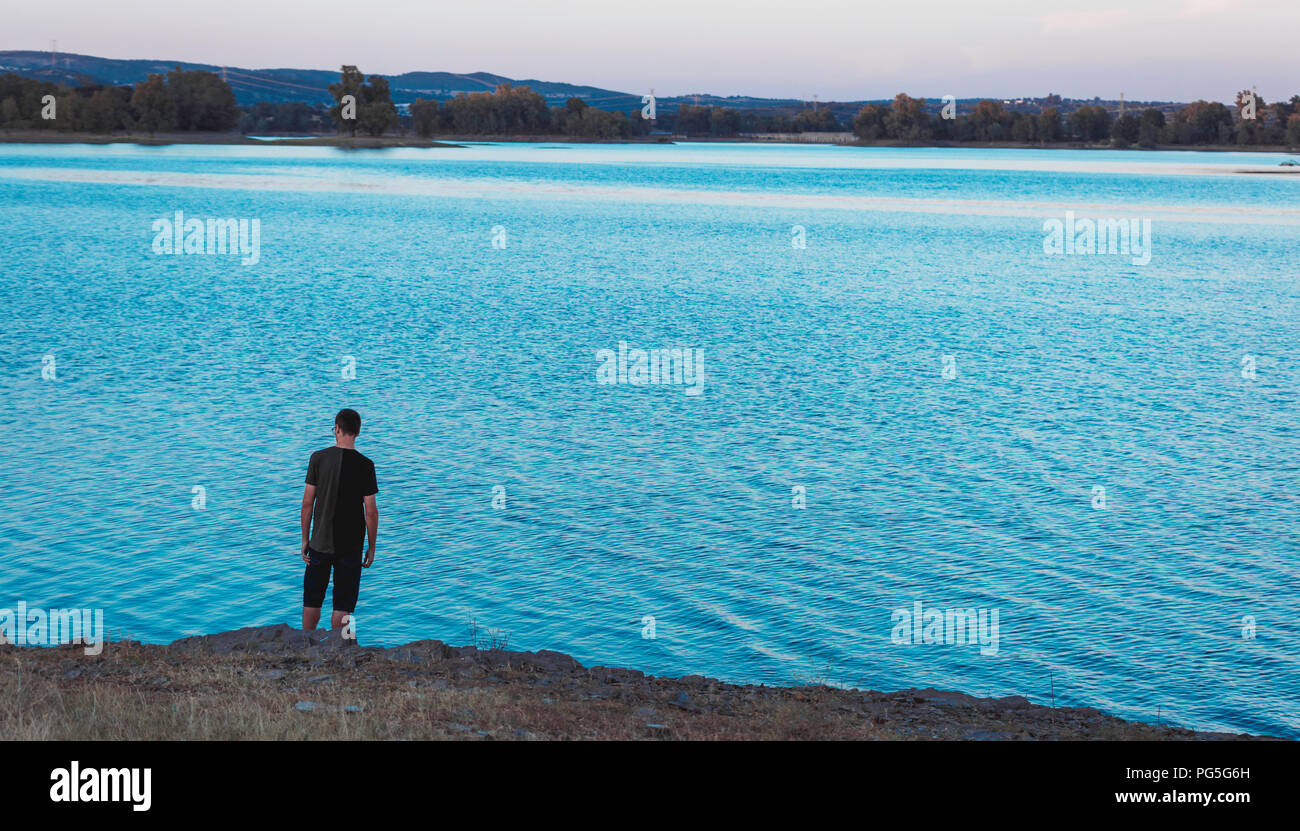 Junge Mann stand und zurück mit legere Kleidung, zurück an den Ufern des Blauen Sees bei Sonnenuntergang, mit Eukalyptus Wald im Hintergrund, Spanien Stockfoto