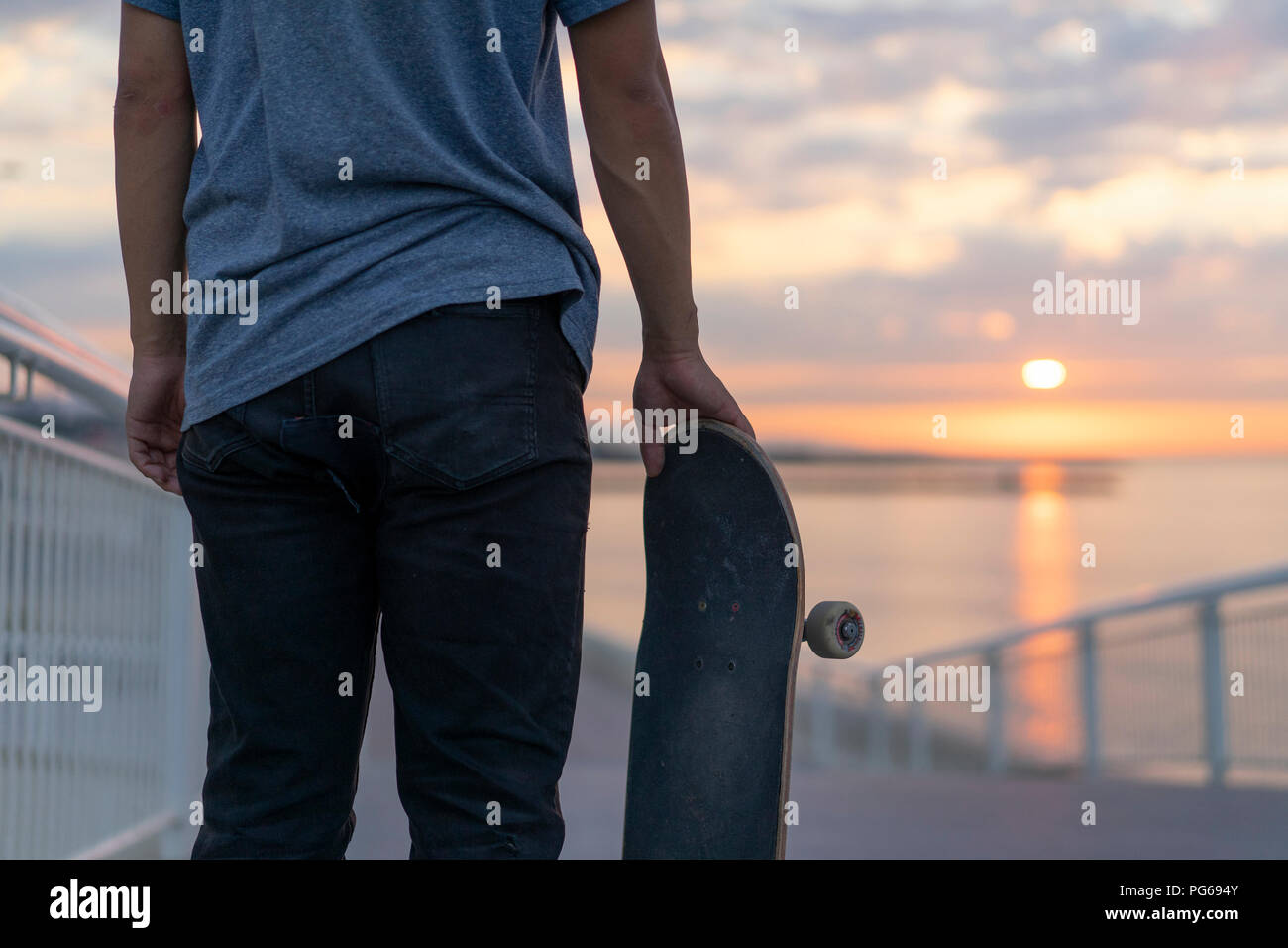 Junger Mann mit Skateboard am Strand bei Sonnenaufgang, Rückansicht Stockfoto