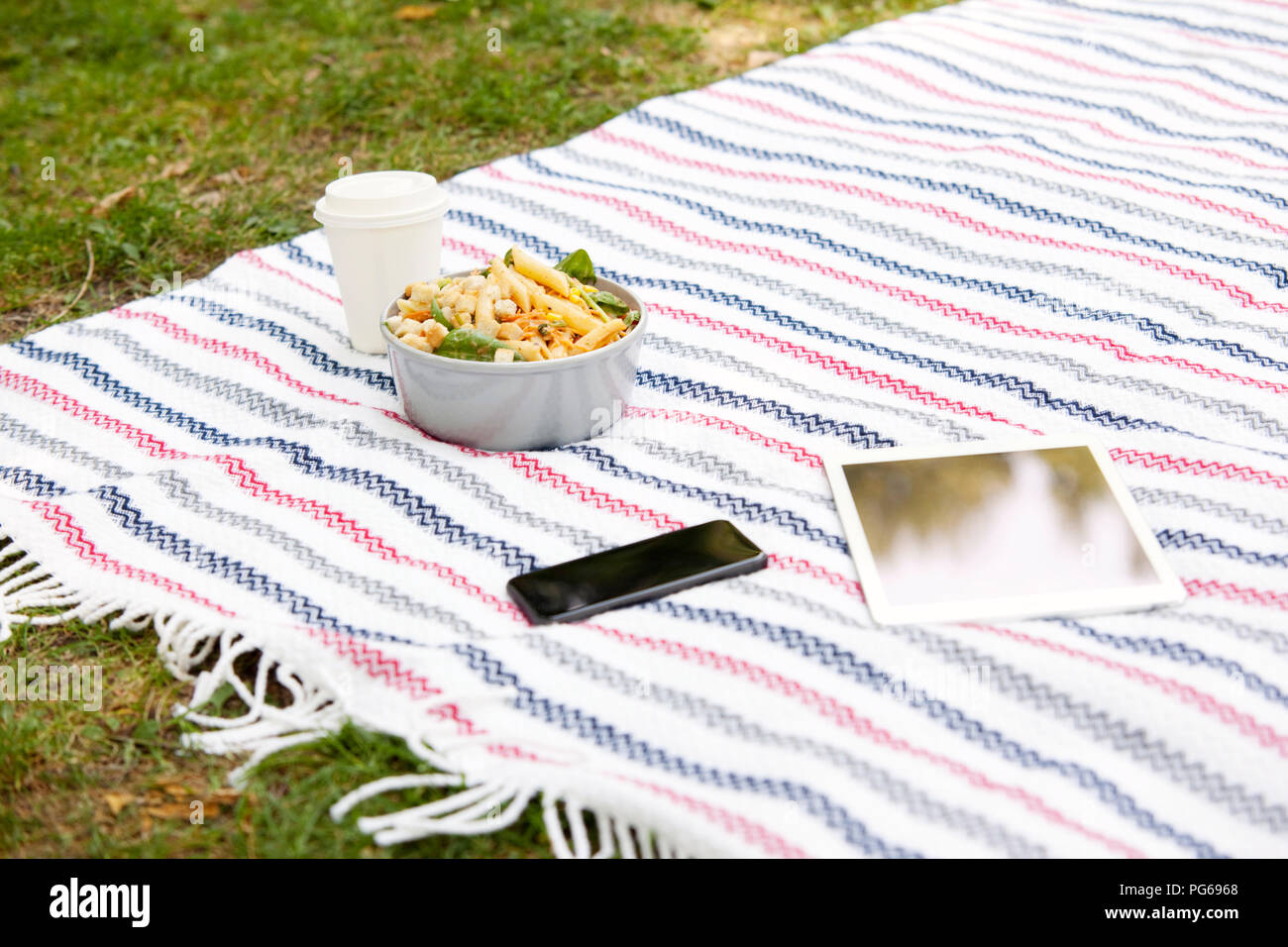 Handy, Tablet, Kaffee trinken gehen und eine Schüssel Nudelsalat auf Decke in einem Park Stockfoto