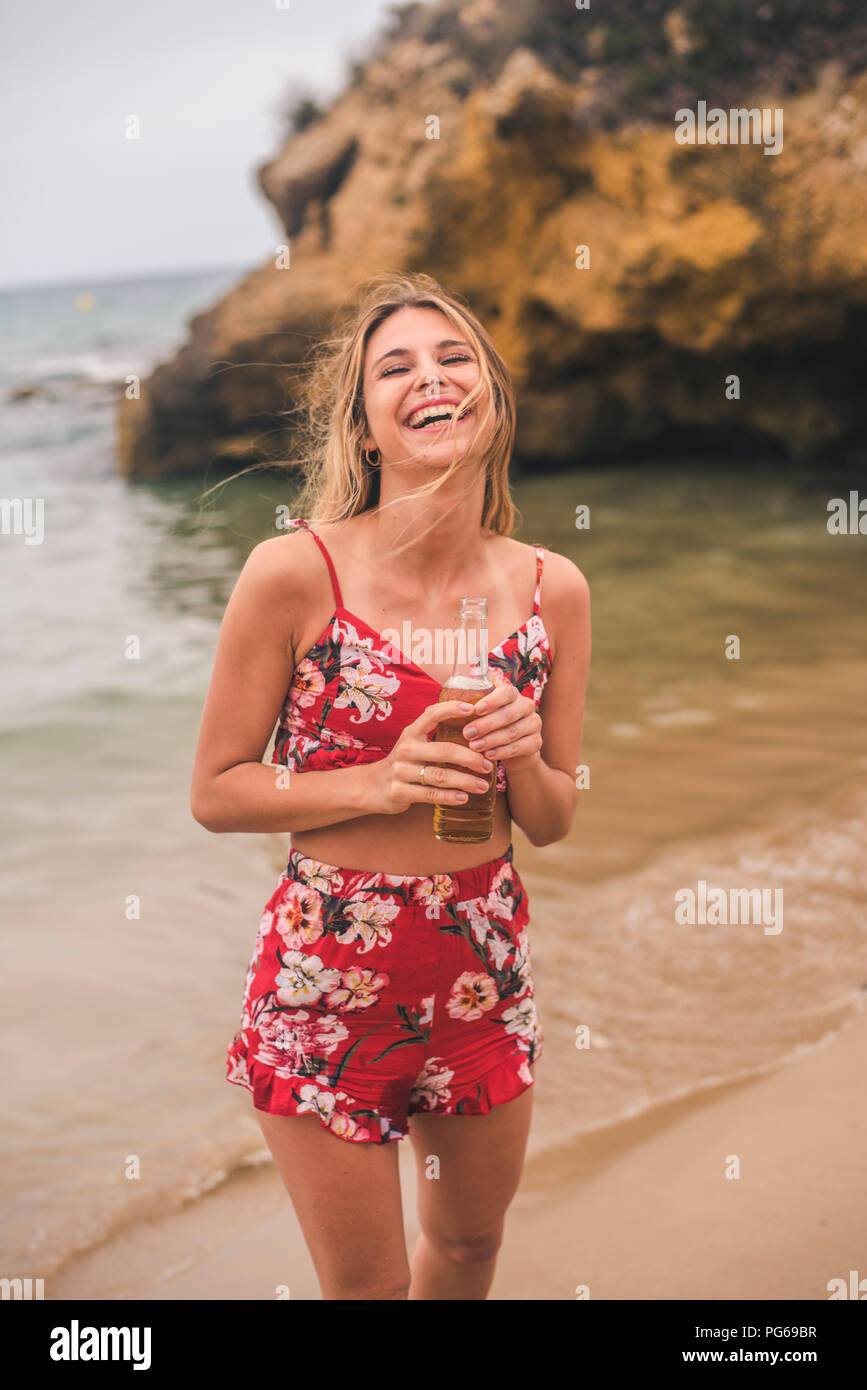 Porträt der glückliche junge Frau, die mit einem Bier am Strand Stockfoto