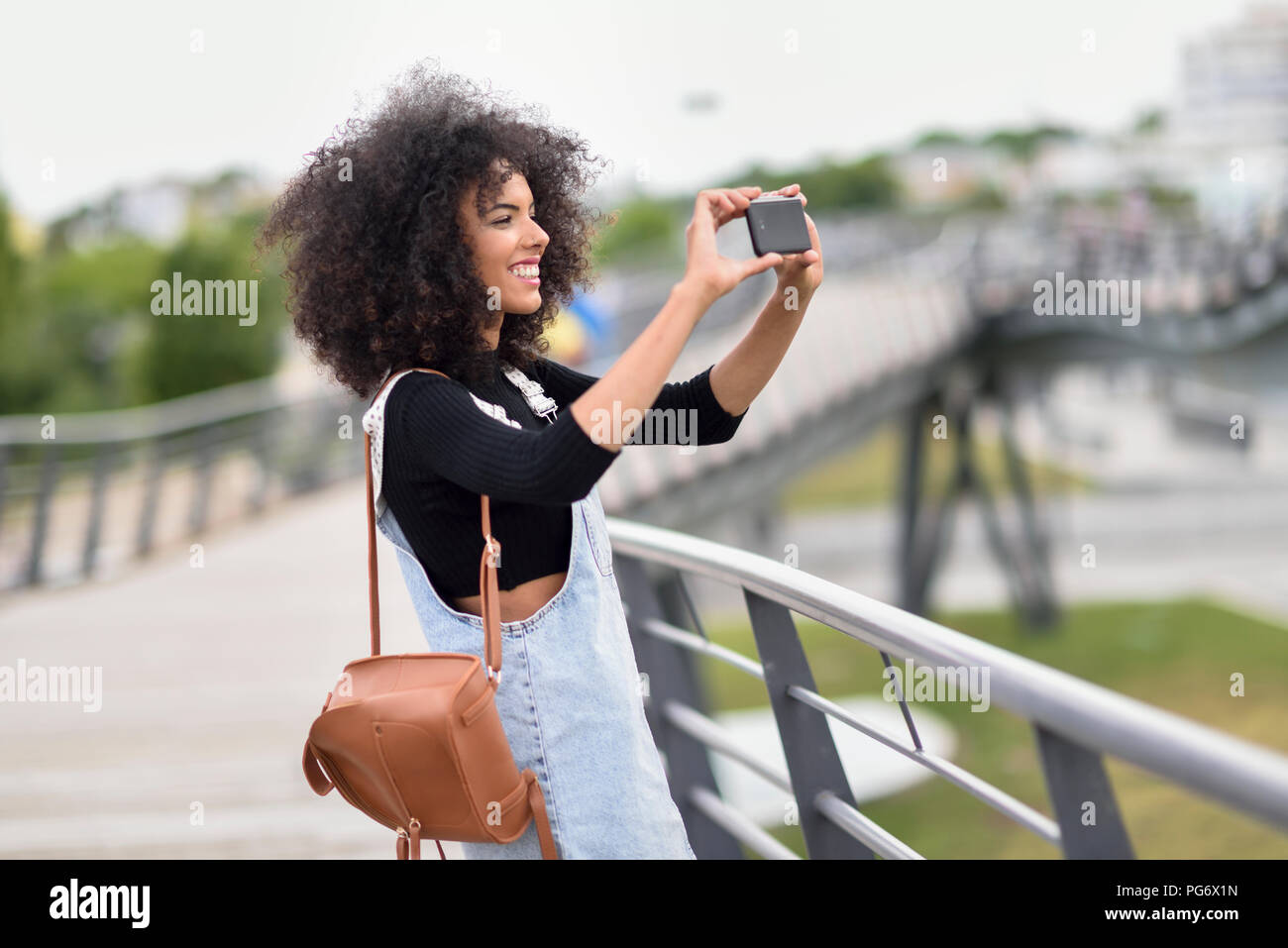 Lächelnde junge Frau mit braunem Leder Rucksack stand auf einer Brücke, um Fotos mit Smartphone Stockfoto