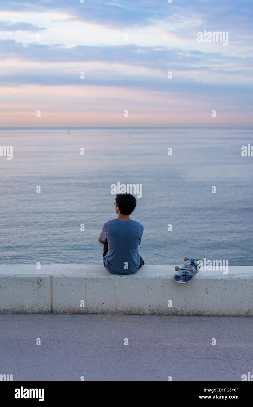 Junge chinesische Mann mit Skateboard sitzen auf Wand am Strand Stockfoto