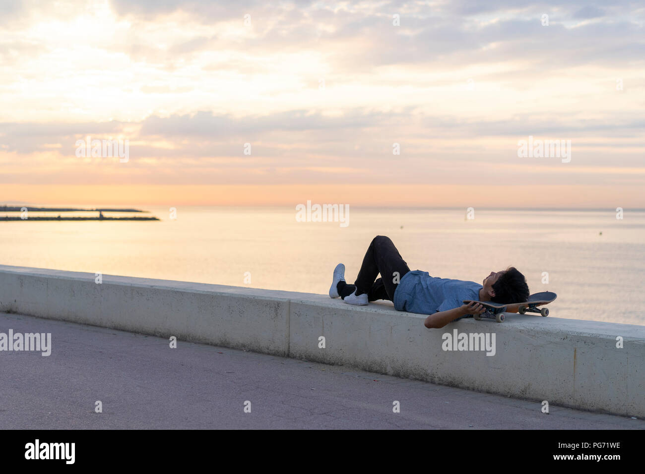 Junge chinesische Mann mit Skateboard liegend an der Wall am Strand Stockfoto