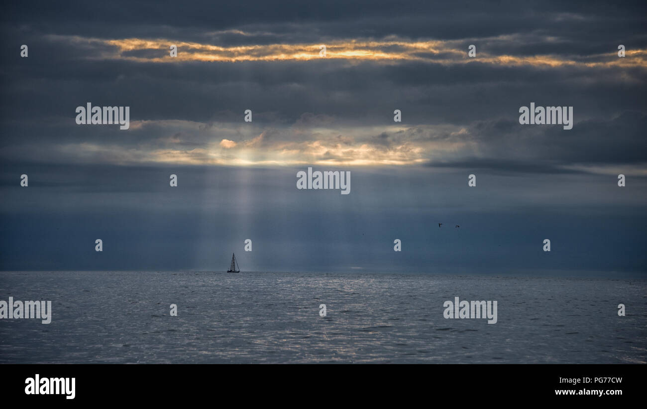 Yachtcharter am Horizont mit Wellen von Dawn Licht durch dramatische Wolken brechen Stockfoto