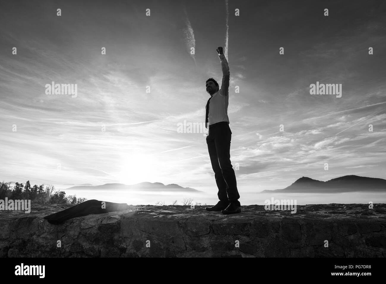 Geschäftsmann Holding aloft seine Faust bei Sonnenaufgang seinen Erfolg und Erfolg feiern, als er auf eine Mauer mit Misty Mountain Szene steht eine Silhouette Stockfoto