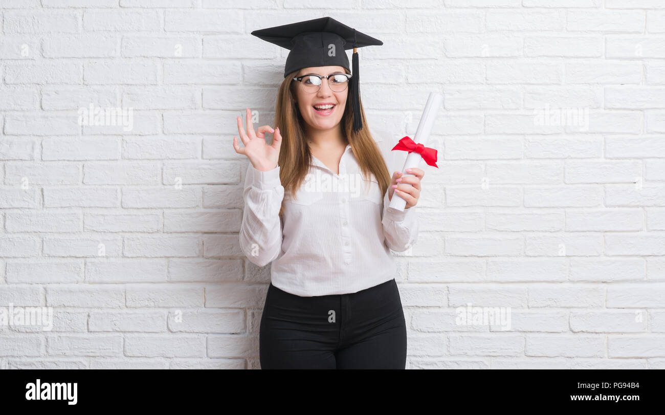 Junge erwachsene Frau über weiße Mauer tragen Absolvent cap Holding Grad tun ok Zeichen mit den Fingern, ausgezeichnete Symbol Stockfoto