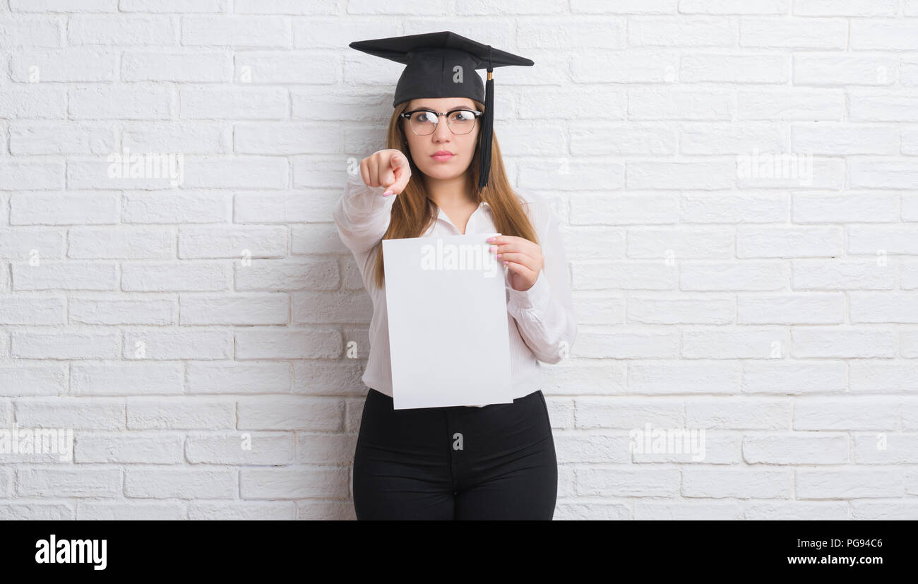 Junge erwachsene Frau über weiße Mauer tragen Absolvent cap Holding grad mit dem Finger an die Kamera und an Sie gerichtet, Handzeichen, positiv und con Stockfoto