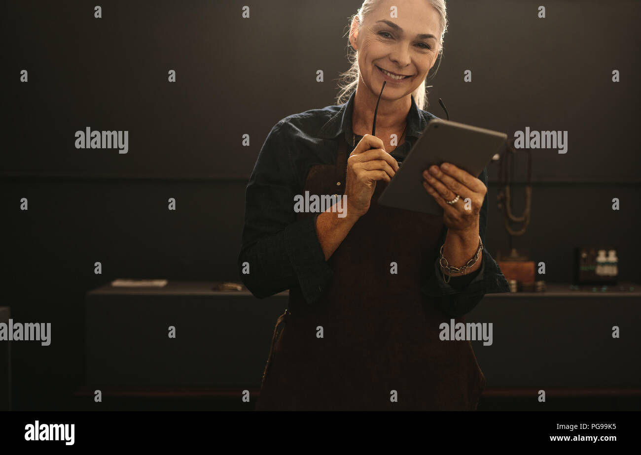 Portrait von lächelnden älteren Frau mit Schürze Holding digital Tablet in ihrem Schmuck Werkstatt. Lächelnd reife weibliche Goldschmied mit Tablet-PC. Stockfoto