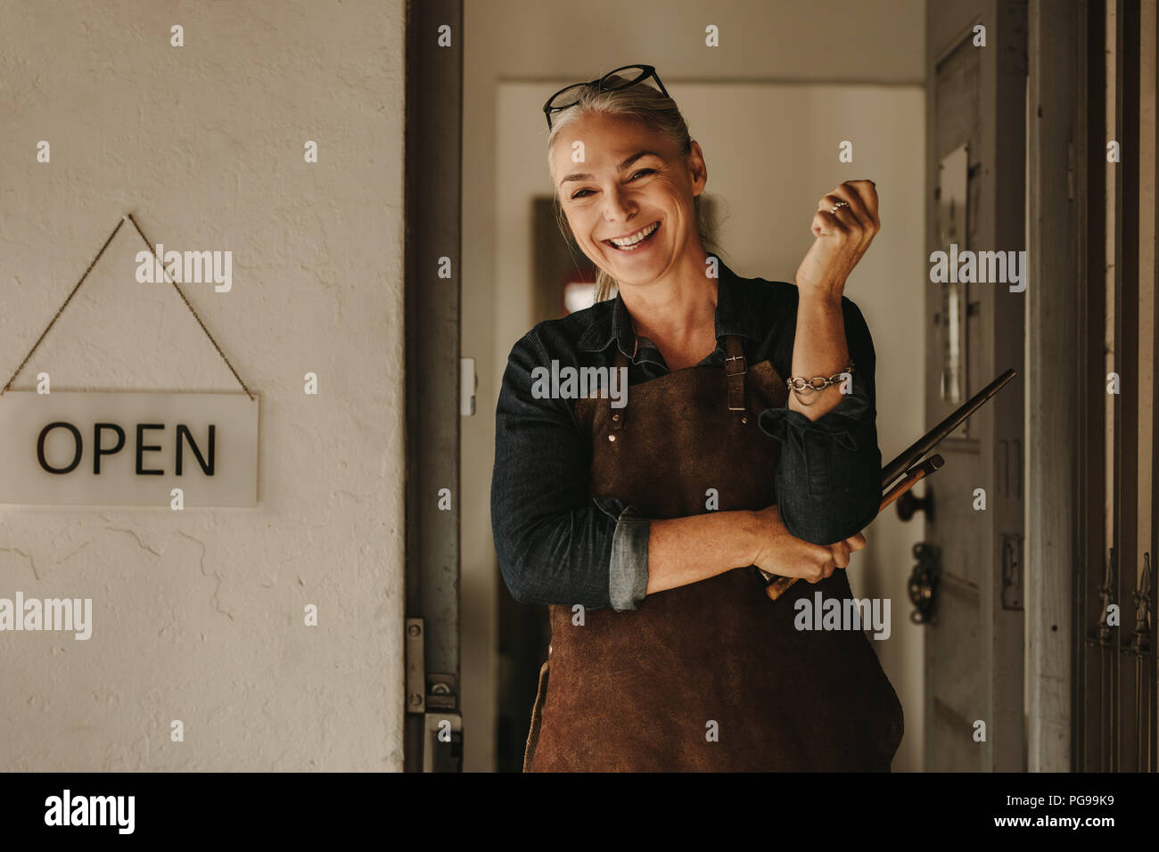 Portrait von fröhlichen älteren weiblichen Juwelier stehend an Workshop Tür. Frau Schmuckhersteller in Schürze können Werkzeuge in der Werkstatt Eingang. Stockfoto