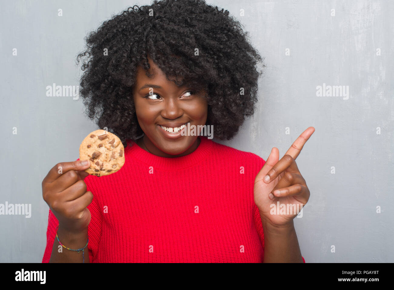 Junge afrikanische amerikanische Frau über Graues grunge Wand essen Chocolate Chip cooky Sehr zufrieden zeigt mit Finger- und an der Seite Stockfoto