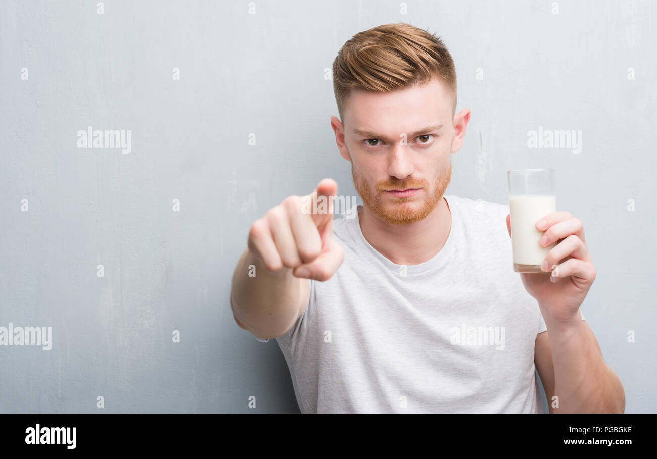 Junge rothaarige Mann über Grau grunge Wand trinkt ein Glas Milch mit dem Finger an die Kamera und an Sie gerichtet, Handzeichen, positiv und zuversichtlich gest Stockfoto