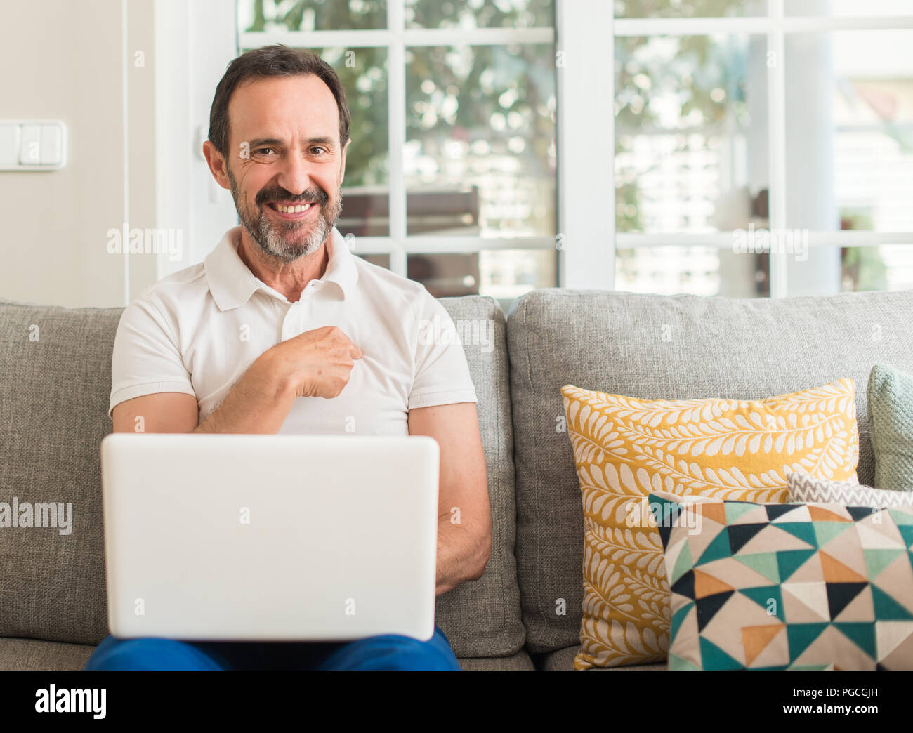 Mittleres alter Mann mit Laptop am Sofa mit Überraschung Gesicht zeigenden Finger zu sich selbst Stockfoto