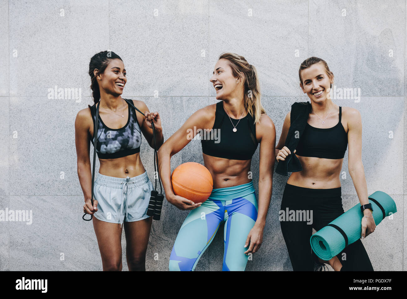 Glückliche Frauen in Fitnesskleidung, die sich entspannen und Spaß haben, an einer Wand zu stehen. Fitness-Frauen tragen Seilbasketball und Trainingsmatte Standi Stockfoto