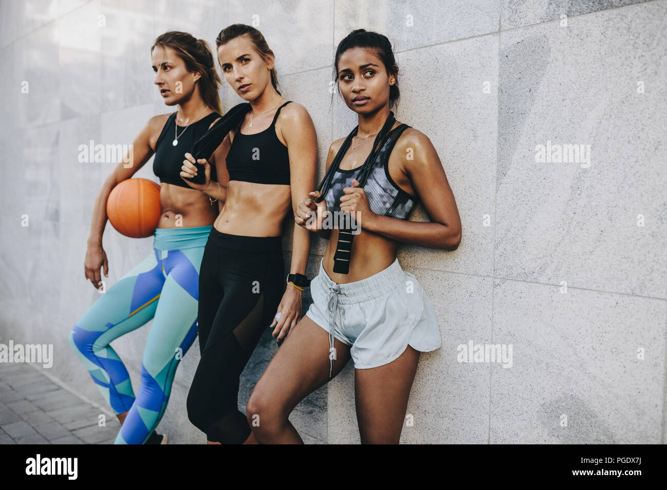Fitness-Frauen, die sich nach dem Workout entspannen und an der Wand stehen. Drei weibliche Athleten stehen mit Seilspringen und Basketball. Stockfoto