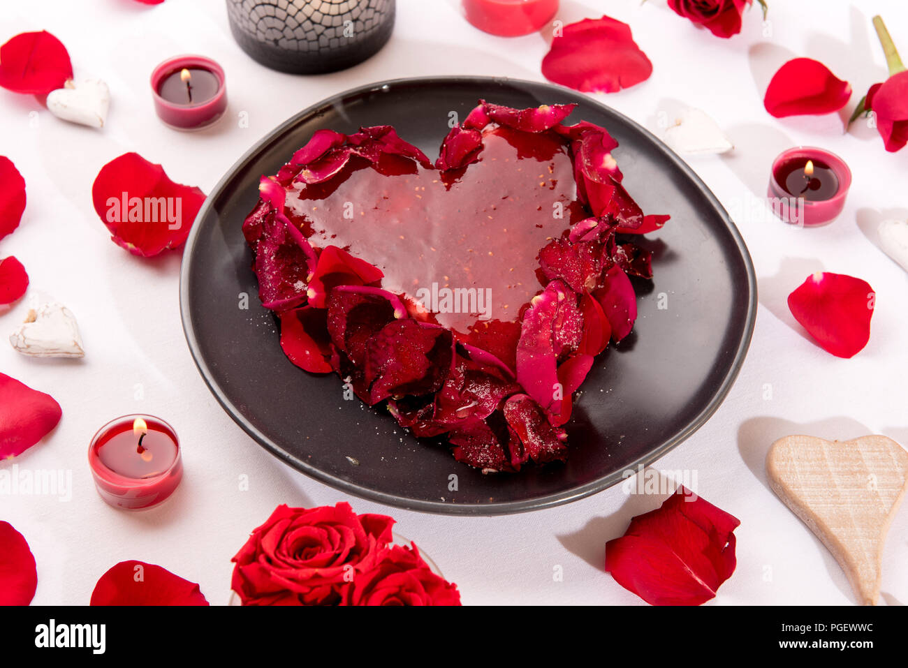 Romantische herzförmige rote Kuchen mit Rosenblättern auf einem weißen Tisch mit Kerzen serviert, Rosenblättern und Herzen für ein Jubiläum oder Valentin Stockfoto