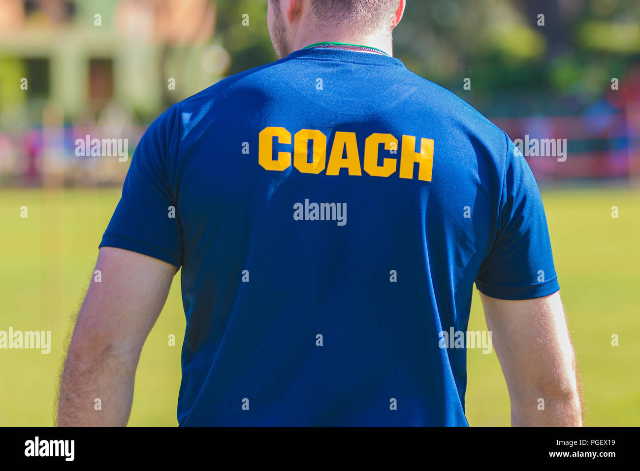 Sport Training Trainer in blaues Hemd mit gelber Trainer Text auf der Rückseite, stehend im Freien an einer Schule Fußball abgelegt Stockfoto