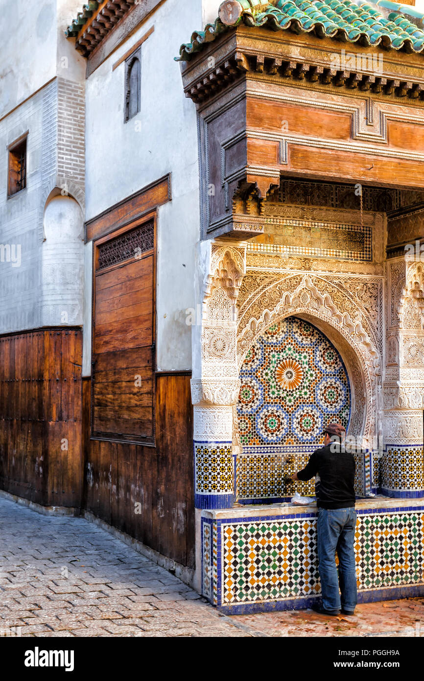 Marokko Mann wäscht seine Hände an ein schönes Mosaik Fliesen- öffentlichen Brunnen und Waschbecken auf einer Straße mit Kopfsteinpflaster im Fes Medina. Stockfoto