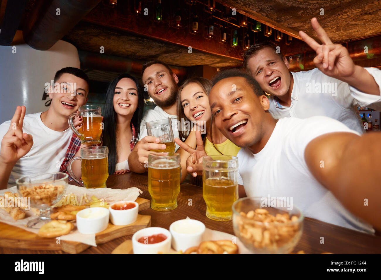 Unternehmen, die glückliche Freunde tragen in Freizeitkleidung, Selbstportrait mit friedenszeichen Geste und trinken Bier, essen Snacks an der Bar. Vier Jungs ein Stockfoto