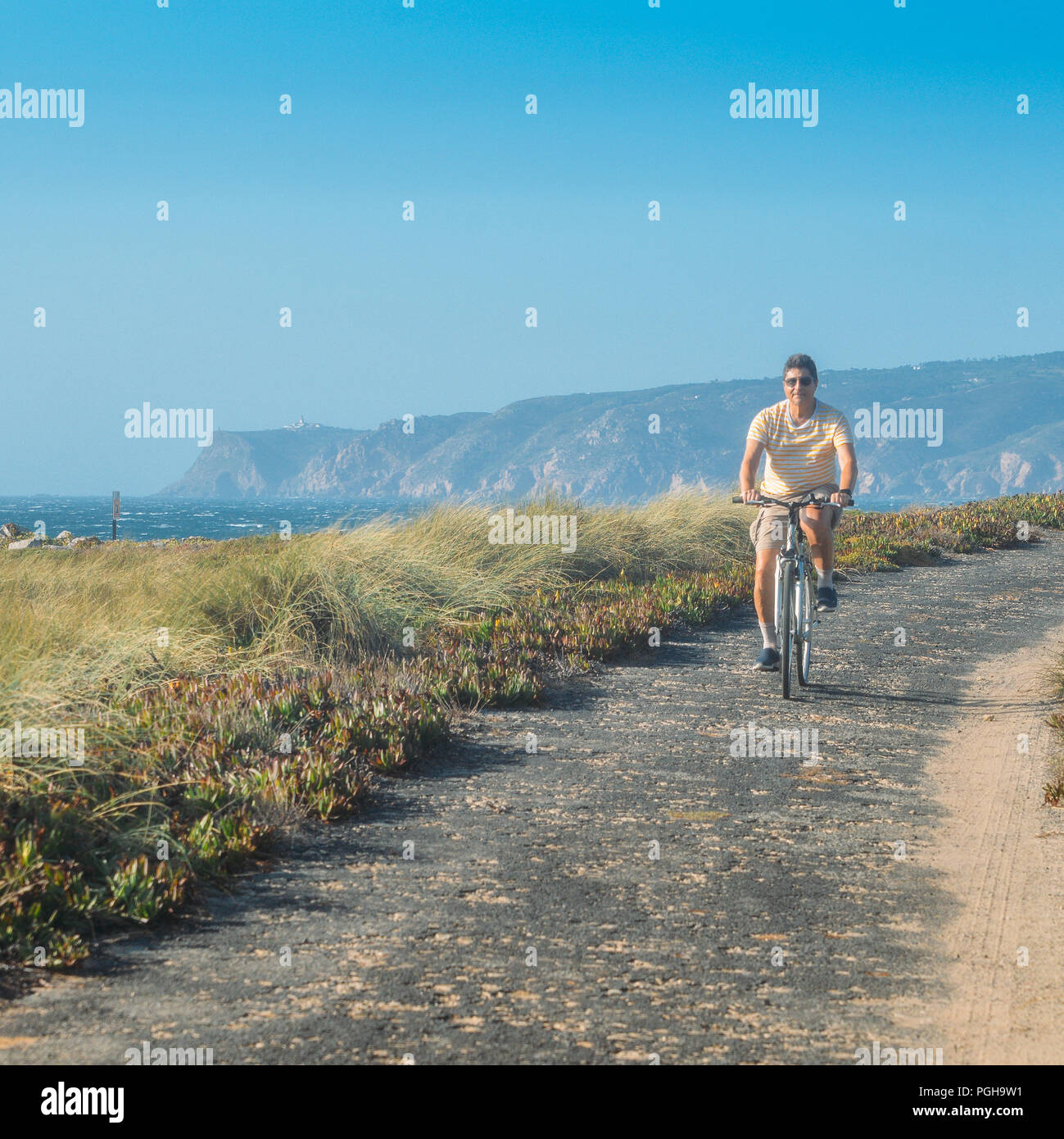 Reifer Mann (55-60) Zyklen entlang ein holpriger Asphalt Weg mit Blick auf den Praia do Guincho und Roca do Cabo in der Nähe von Cascais, Portugal während eines windigen Sommer Tag Stockfoto