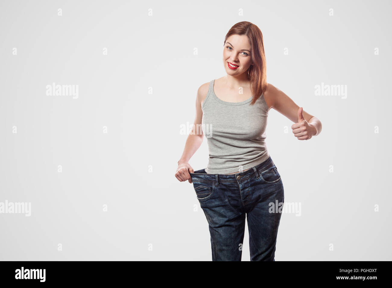 Portrait von glücklich Schöne schlanke Taille der jungen Frau in big Jeans und grau top zeigen erfolgreiche Gewichtsabnahme, Innen, studio Shot, auf Licht isoliert Stockfoto