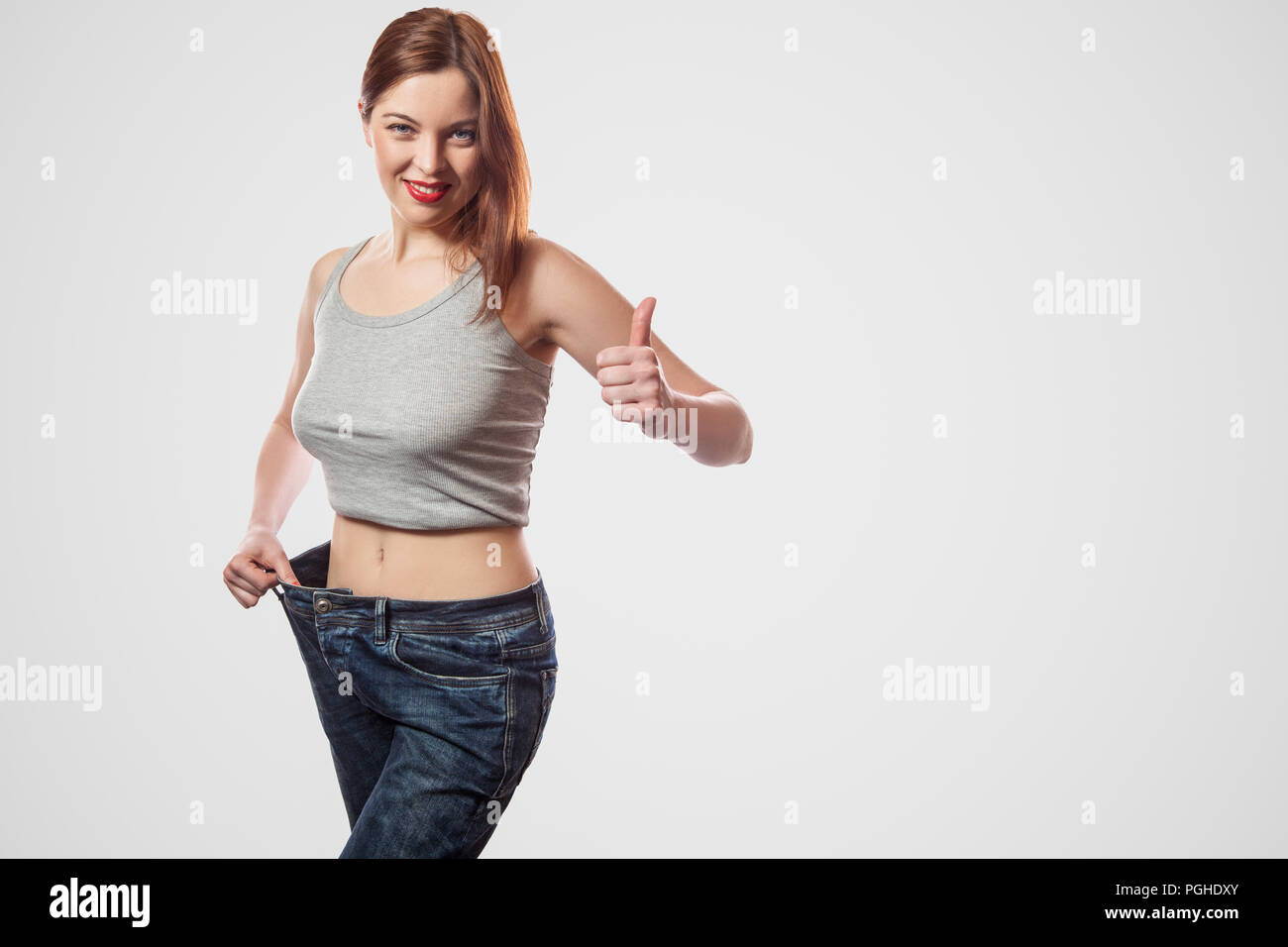 Portrait von glücklich Schöne schlanke Taille der jungen Frau, die in Jeans und grau top zeigen erfolgreiche Gewichtsabnahme, Innen, studio Shot, isoliert Stockfoto