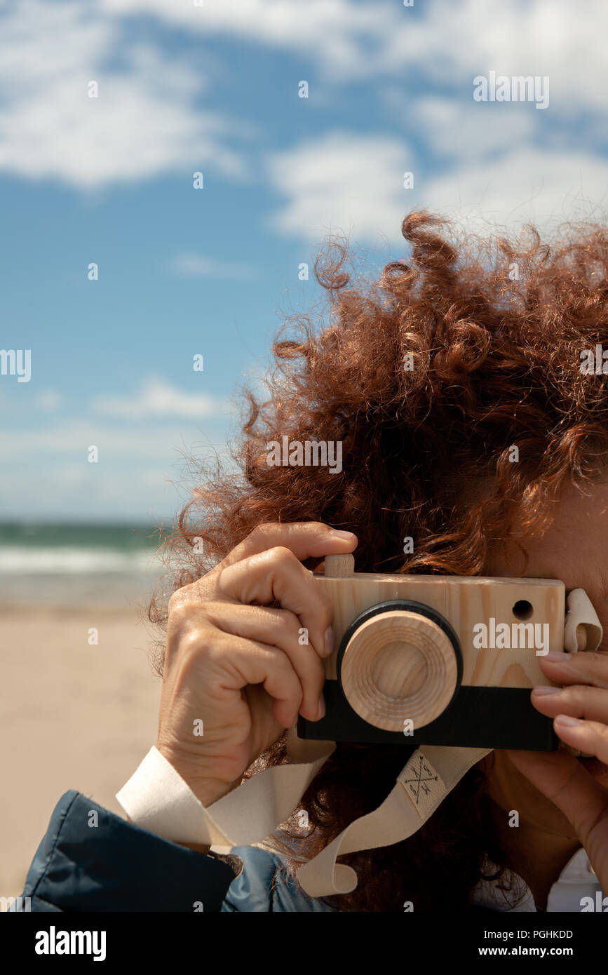 Rot lockiges Haar schöne junge Frau Porträt hält eine hölzerne Spielzeug Foto Kamera vor sandigen Strand Hintergrund als Fotografie-Konzept. Stockfoto