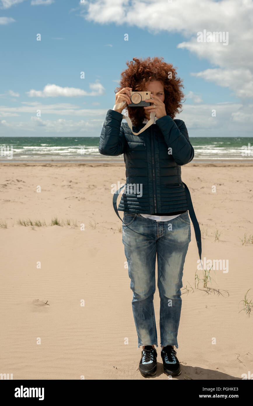 Schöne junge Frau mit dem lockigen Haar, die im Besitz einer Holzspielzeug Foto Kamera auf leeren Sandstrand Hintergrund. Spaß Fotografie Konzept. Stockfoto