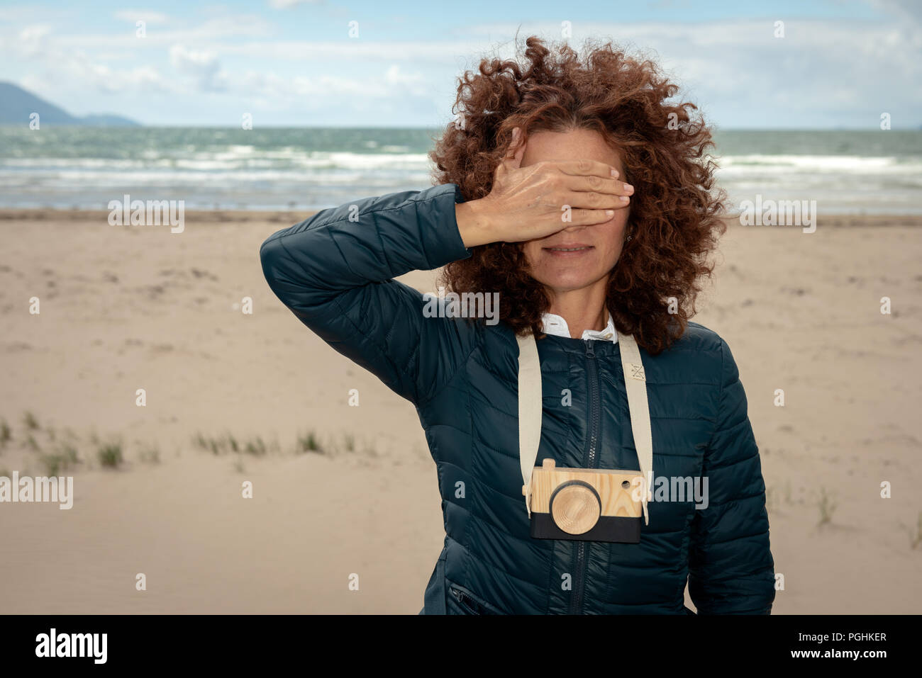 Junge, wunderschöne Frau mit roten Locken, die mit einer hölzernen Spielzeug-Fotokamera am leeren Strand posiert und dabei ihre Augen mit der Hand versteckt Stockfoto