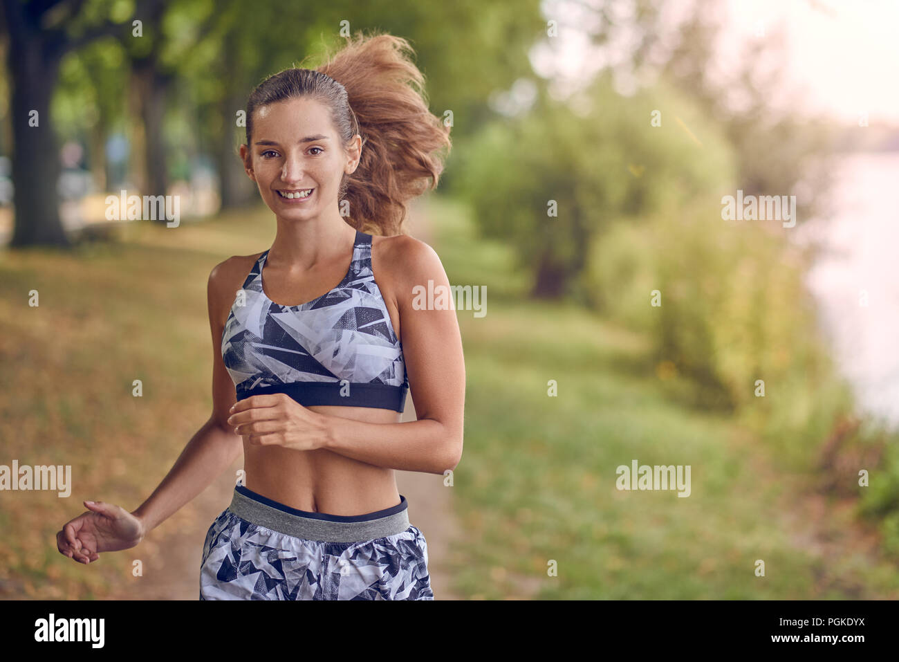 Fit gesund athletische Frau Joggen an einem Flussufer glücklich lächelnd mit Ihren langen Haare hinter ihr fliegen wie Sie nähert sich die Kamera Stockfoto