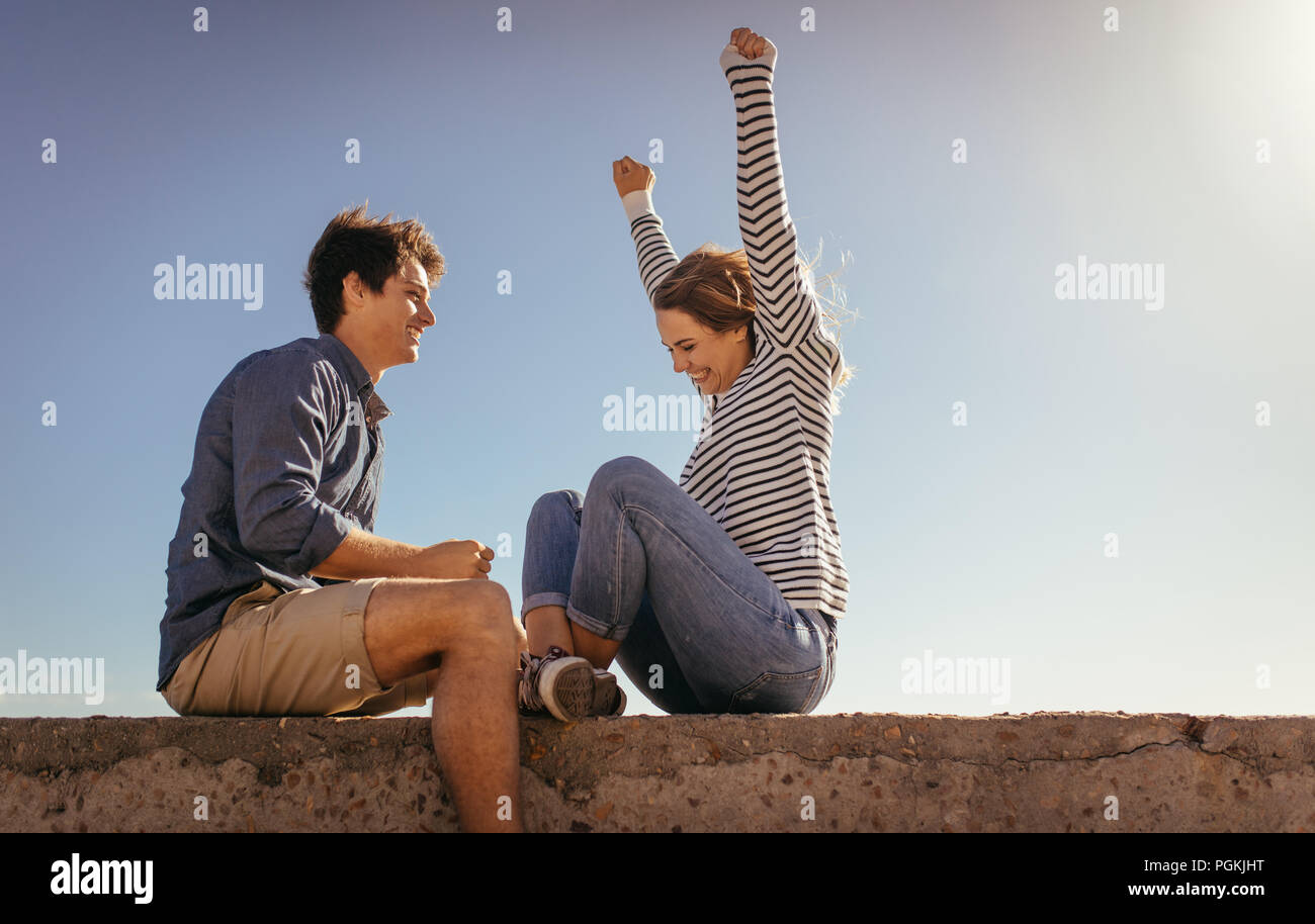 Aufgeregt Frau mit erhobenen Händen sitzen auf einem Sea Wall mit ihrem Freund. Paar sitzt auf einem Ferienhäuser sitzen auf eine Betonwand an einem sonnigen Tag. Stockfoto