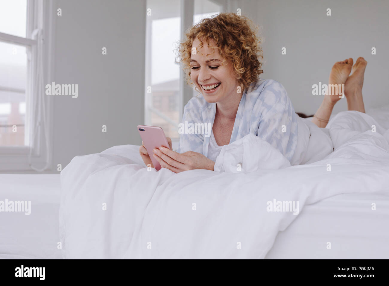 Glückliche Frau liegend auf Bett prüfen ihr Handy. Frau mit Ihrem Mobiltelefon im Bett nach dem Aufwachen am Morgen. Stockfoto