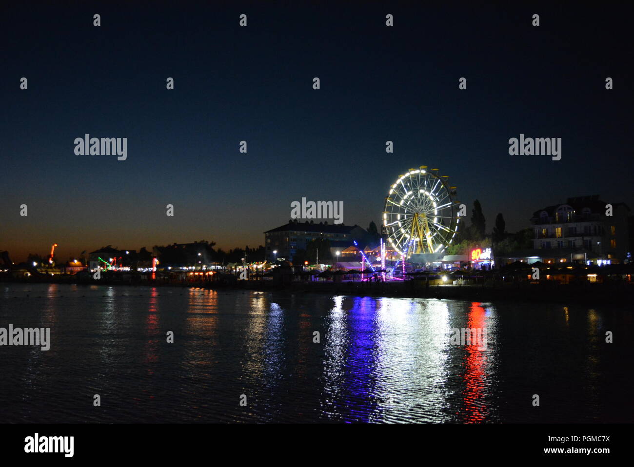 Nachtunterhaltung und Entspannung am Strand des Schwarzen Meeres mit Attraktionen, weißem Riesenrad, Trampolin in einer hellen, blauen, gelben Farbe. Stockfoto