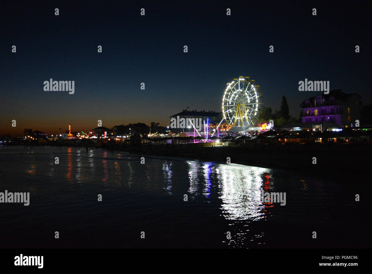 Nachtunterhaltung und Entspannung am Strand des Schwarzen Meeres mit Attraktionen, weißem Riesenrad, Trampolin in einer hellen, blauen, gelben Farbe. Stockfoto