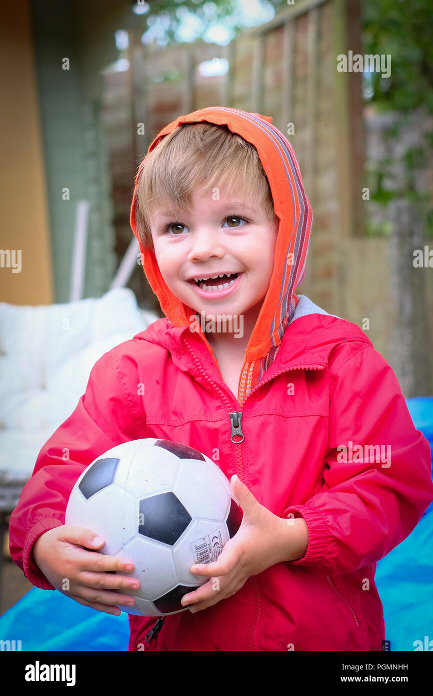 Jungen im Alter von zwei (2) mit roten Mantel und Fußball außerhalb glücklich und lächelnd. Stockfoto