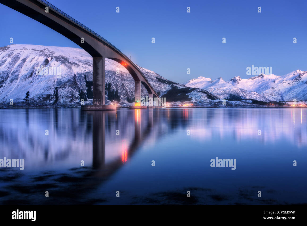 Brücke mit Beleuchtung, schneebedeckte Berge, Dorf und blauer Himmel mit wunderschönen Reflexion im Wasser. Nacht Landschaft mit Brücke, verschneite Felsen re Stockfoto