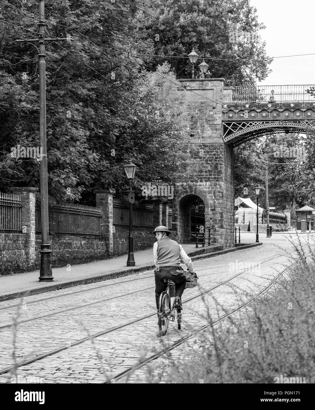 Nostalgisch, schwarz-weiß, Rückansicht eines einsamen Radfahrers aus den 40er Jahren im Blechhelm, der auf einer verlassenen, gepflasterten Straße entlang fährt und zurückblickt. Stockfoto