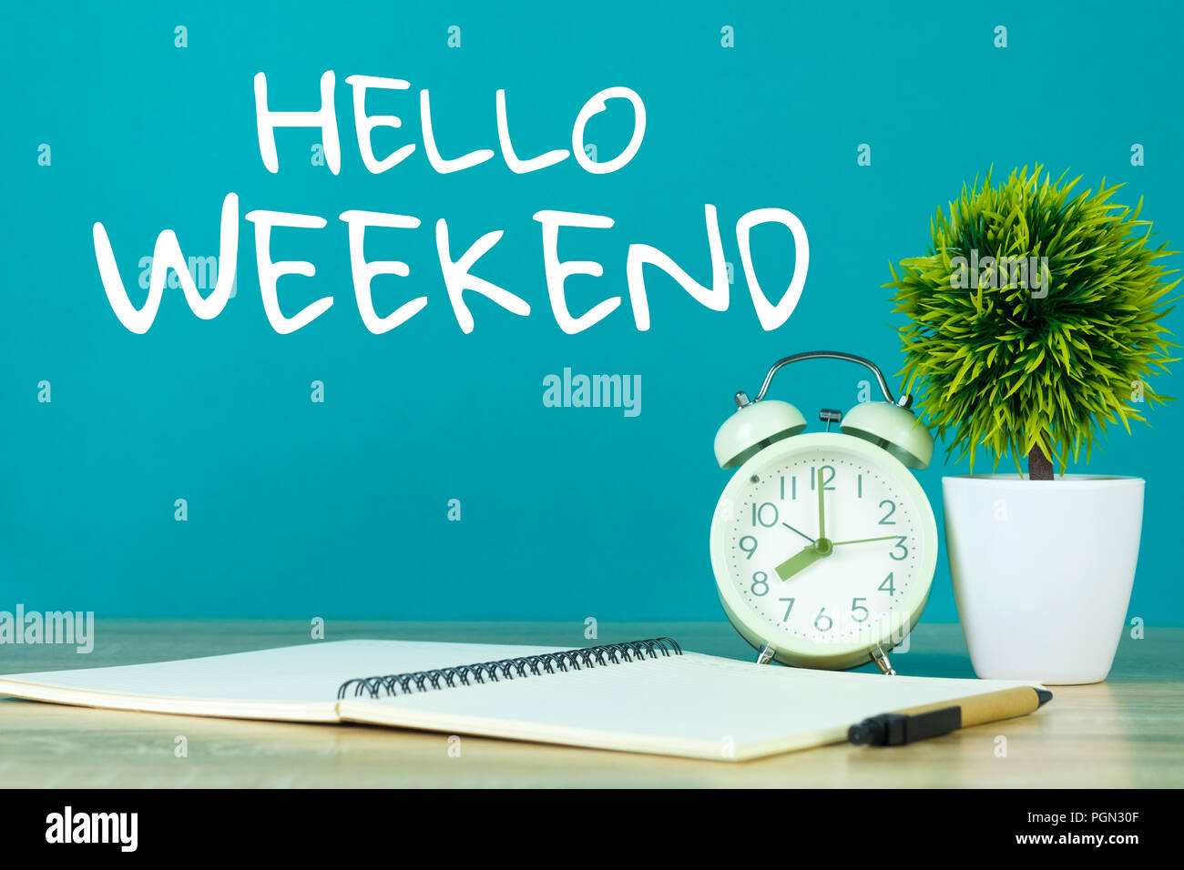 Hallo Wochenende Text auf grünem Hintergrund und kleinen Baum in weiße Vase mit Vintage Wecker und Notebook Papier Stift auf Holztisch. Stockfoto
